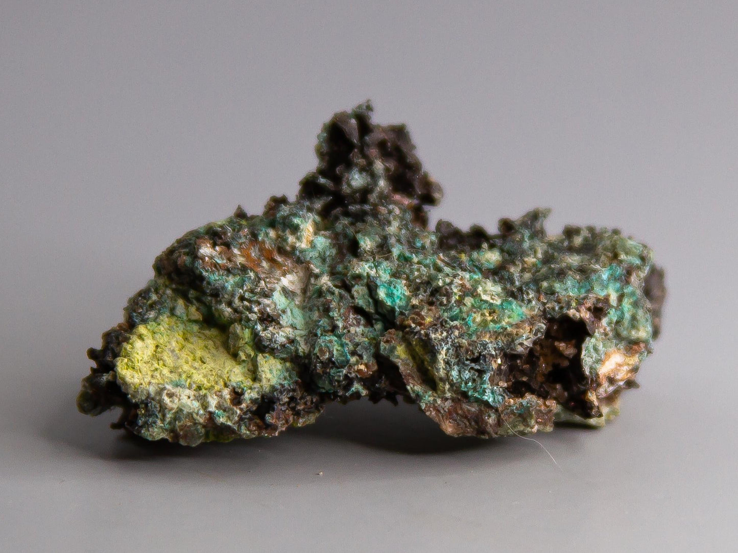 Mineral sample with green and brown colors on a gray background, branded 'Adamantine Minerals'.
