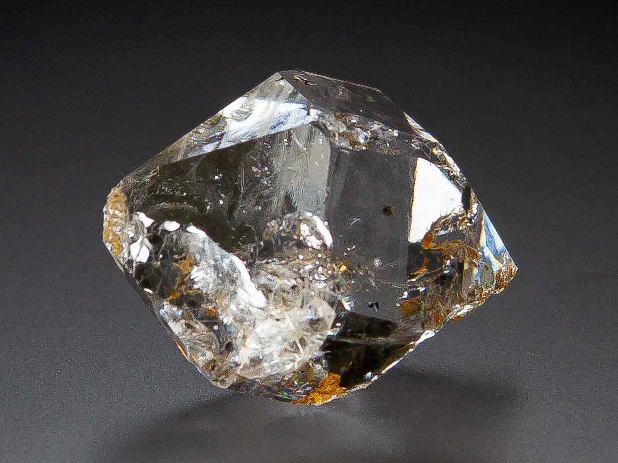 A raw, Herkimer Diamond specimen with visible internal inclusions and small brownish inclusions, photographed on a dark gray background.