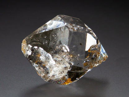 A raw, Herkimer Diamond specimen with visible internal inclusions and small brownish inclusions, photographed on a dark gray background.