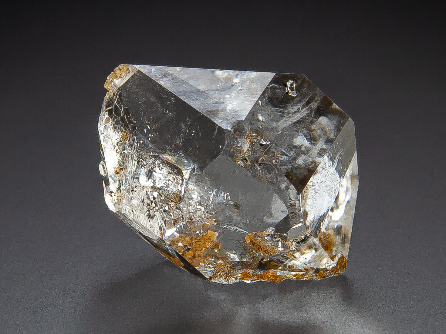 A raw, clear Herkimer Diamond crystal specimen with natural inclusions and small brownish mineral deposits, photographed on a dark gray background.