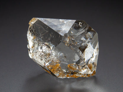 A raw, clear Herkimer Diamond crystal specimen with natural inclusions and small brownish mineral deposits, photographed on a dark gray background.