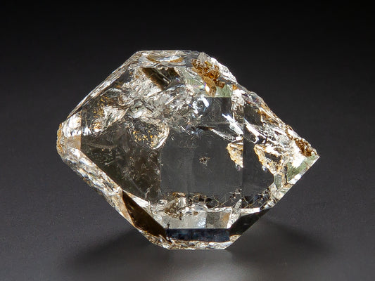 A raw, clear gemstone with natural inclusions and a faceted, geometric shape, photographed on a dark black background.