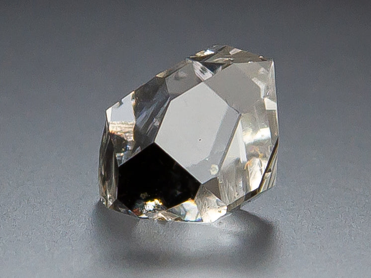 A clear Herkimer Diamond with flat glassy faces, photographed on a smooth gray surface with a soft shadow.