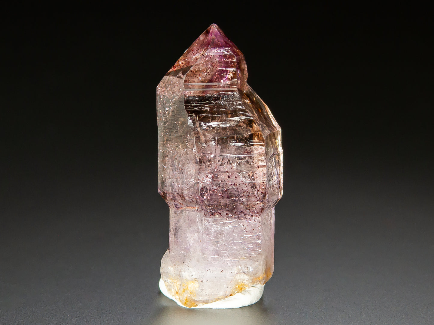 A natural amethyst crystal point specimen with pink and purple internal inclusions, standing upright on a white base against a dark gray background.