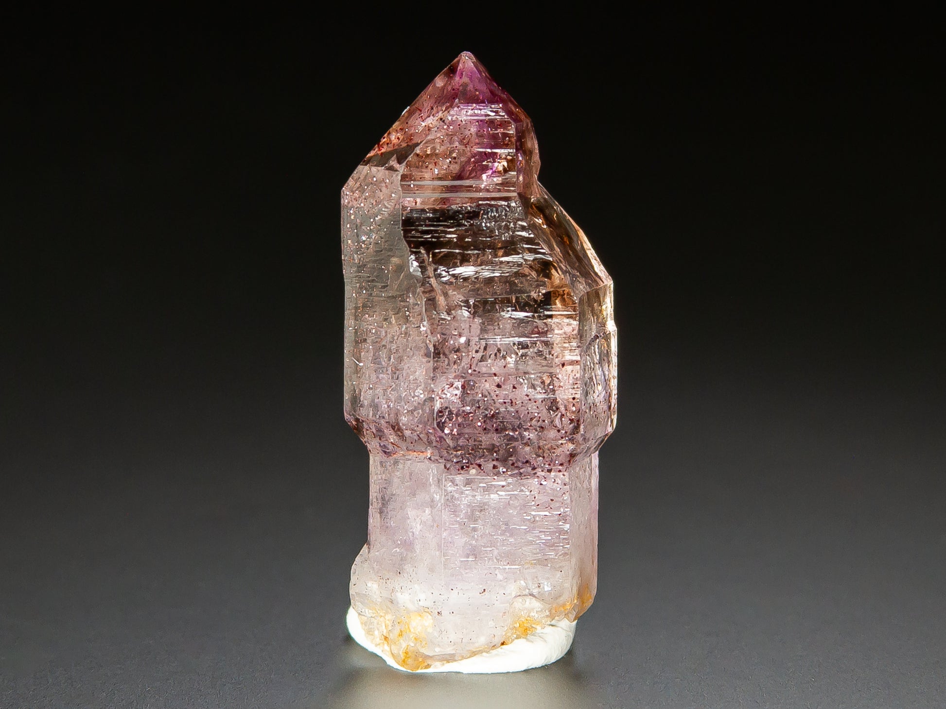 A natural amethyst crystal point specimen with pink and purple internal inclusions, standing upright on a white base against a dark gray background.