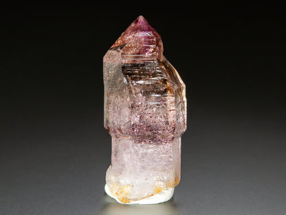 A natural amethyst crystal point specimen with pink and purple internal inclusions, standing upright on a white base against a dark gray background.