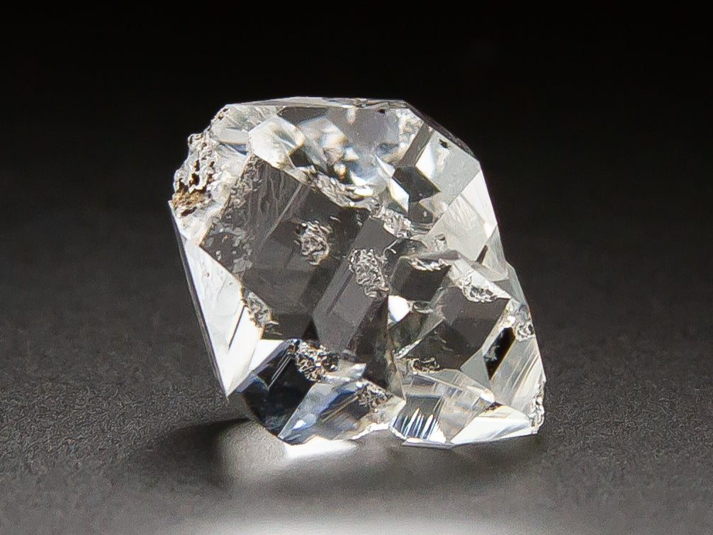 A clear, faceted crystal or gemstone with rough, natural edges, photographed on a dark gray to black background.